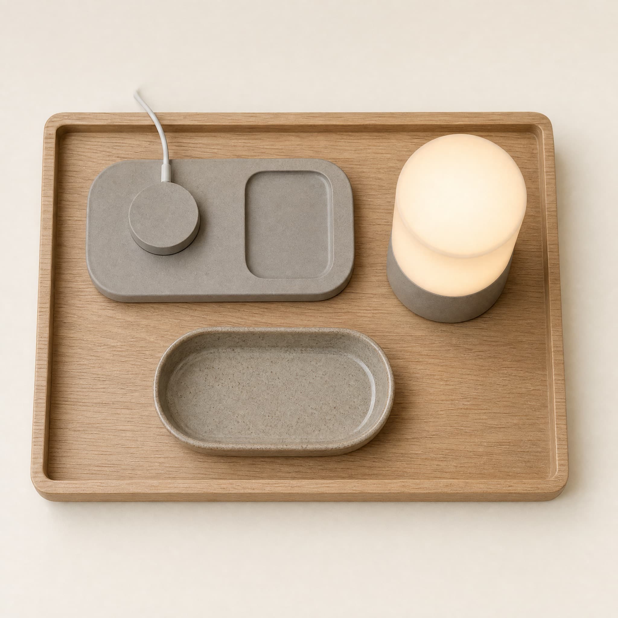 Bedside Core Kit flat lay: oak bedside tray, rounded charging dock, glazed stoneware dish and a compact lamp with a soft warm glow.